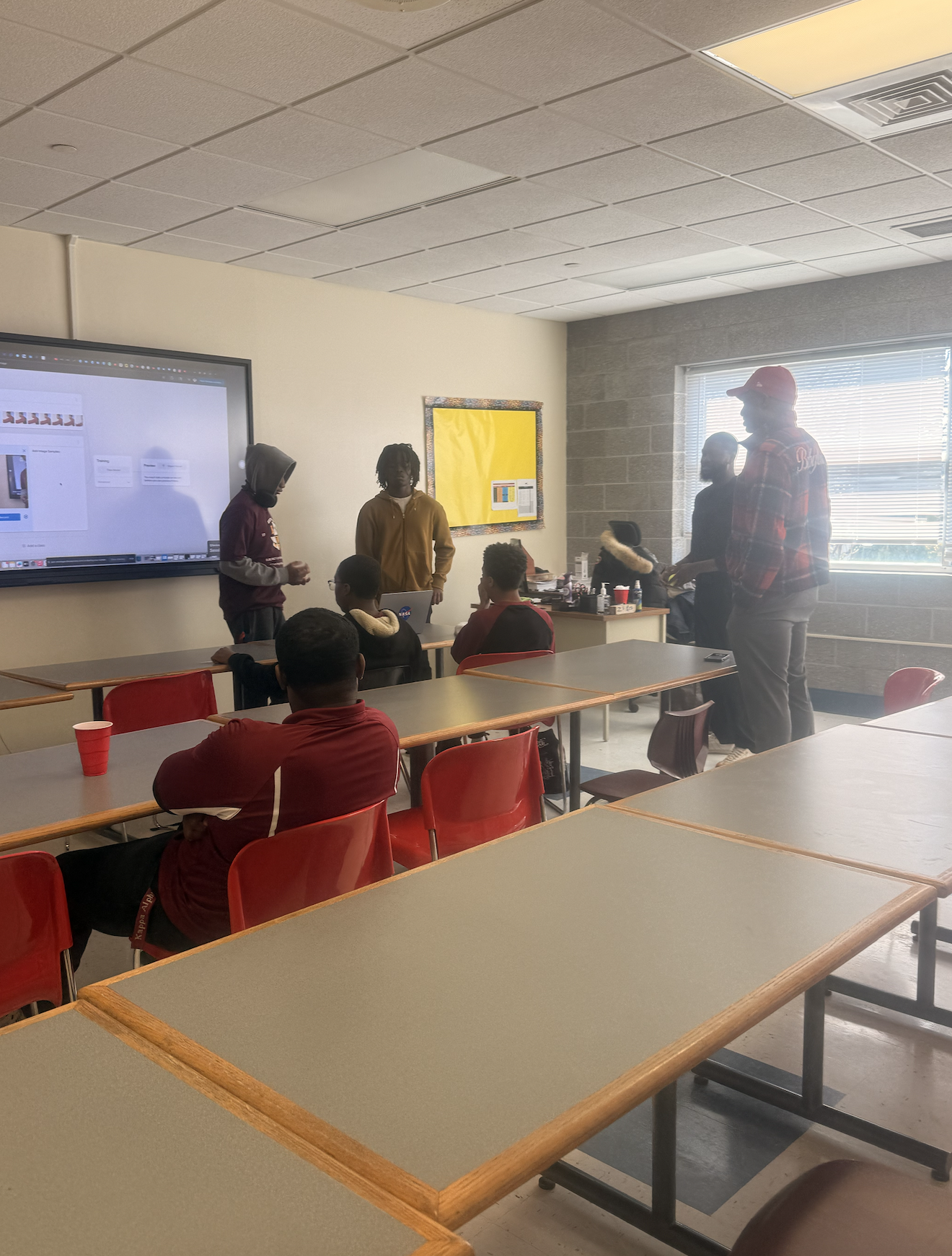 Students and mentors standing at the front of a classroom beside a screen showing an A.I. training tool