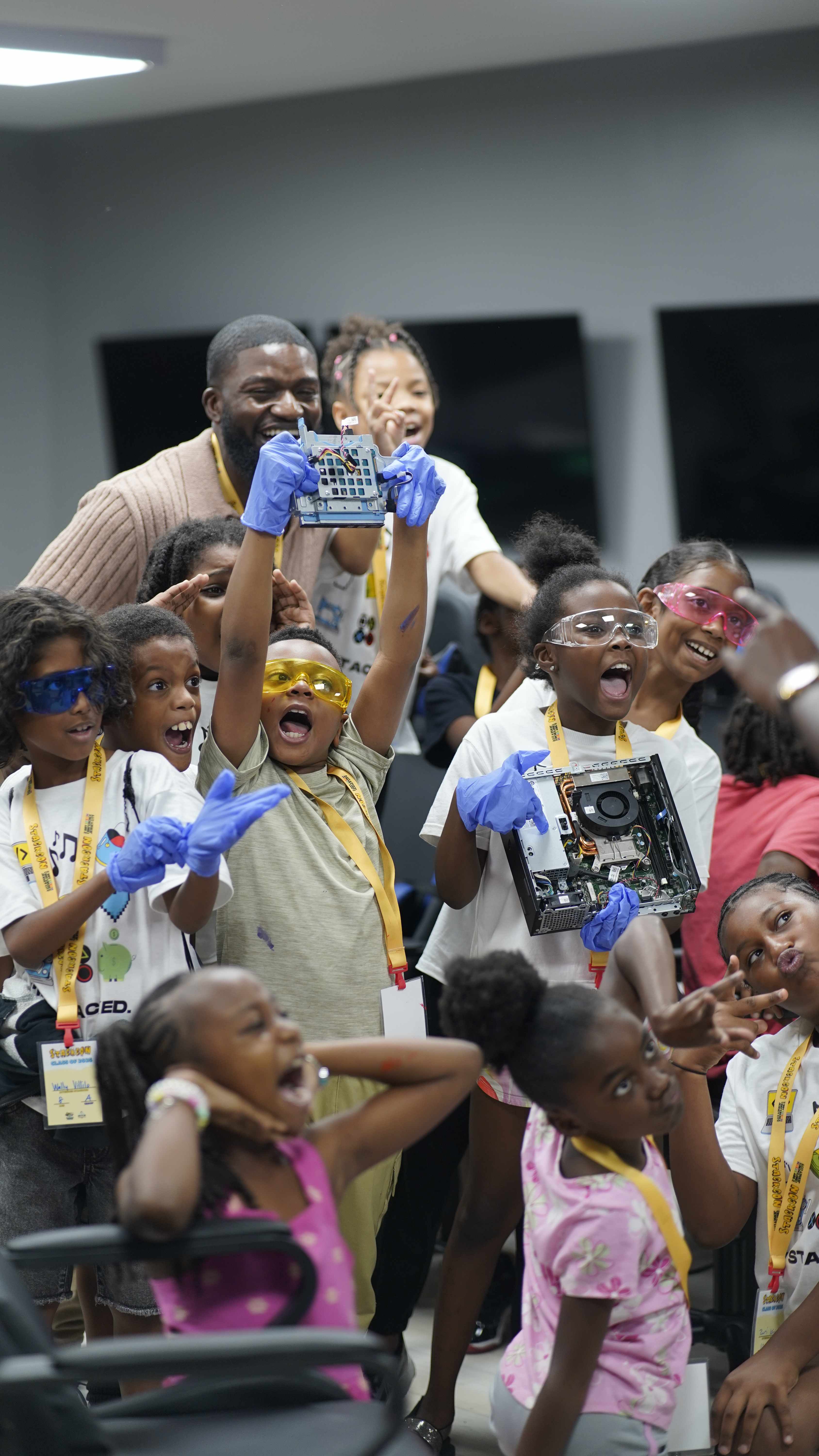 Excited kids in safety goggles and gloves cheering while holding up computer parts they took apart at StackCon